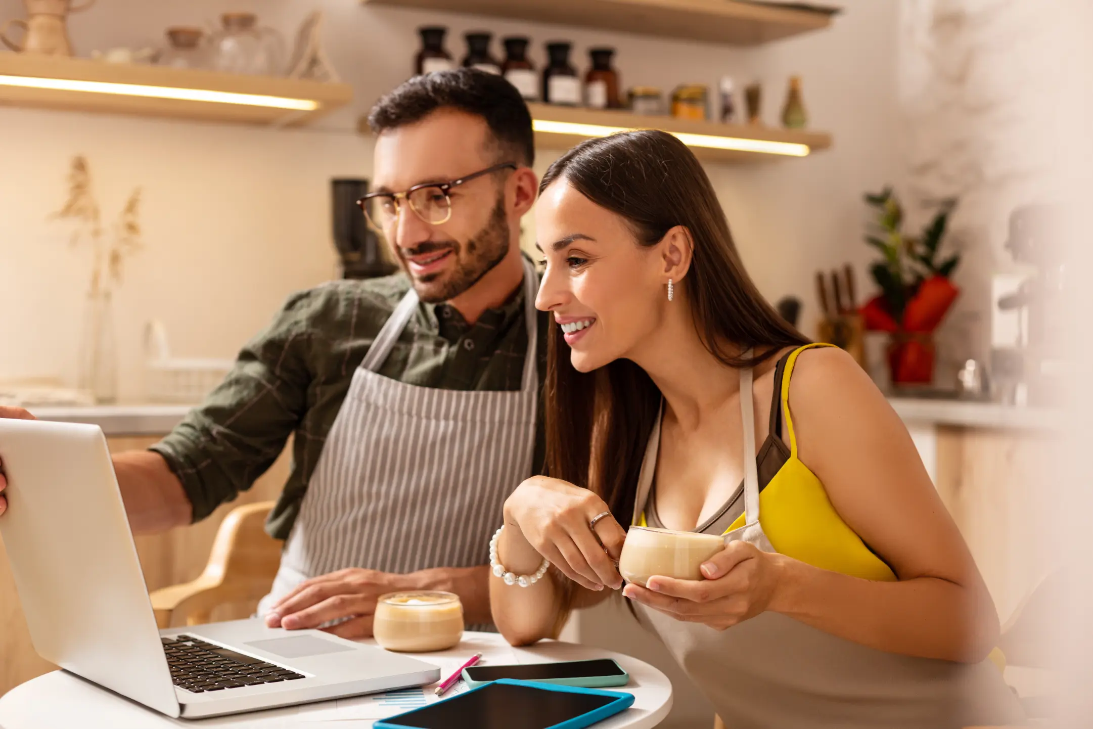 Homem e mulher estão com aventais em uma cozinha e decidem o menu do restaurante no notebook