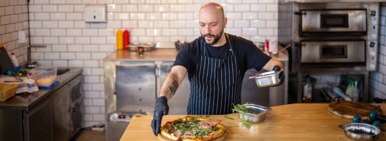 economia 2023: pizzaiolo com avental e luvas na cozinha terminando de rechear uma pizza em cima da bancada