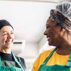 auxiliar de cozinha: duas cozinheiras uma branca e outra negra utilizando avental e touca dentro da cozinha