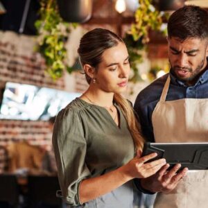 balanced scorecard: empreendedora branca mostrando tablet para funcionário negro que utiliza um avental. Ambos estão dentro do restaurante.