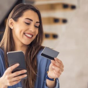 banco digital: mulher sentada e sorrindo em cadeira enquanto olha o celular segurando um cartão de crédito