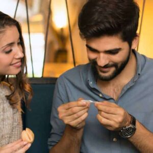 biscoito da sorte: casal de homem e mulher brancos jantando com taças de vinho. A mulher segura um biscoito da sorte e o homem lê a mensagem que veio no dele.