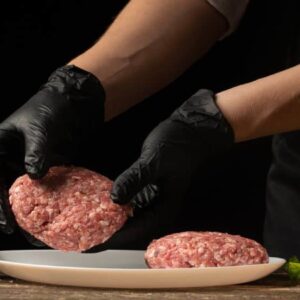 blend de carne: homem utilizando luvas e avental pretos coloca hambúrgueres crus em prato na mesa com vários condimentos, temperos e vegetais