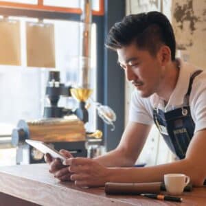 cafeteria: homem barista utilizando avental se apoiando em balcão para olhar tablet