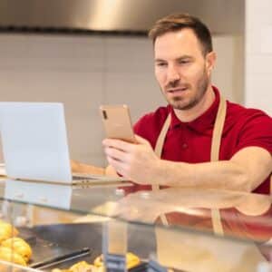 cardápio: homem vestindo avental atrás do balcão de padaria enquanto verifica notebook e celular