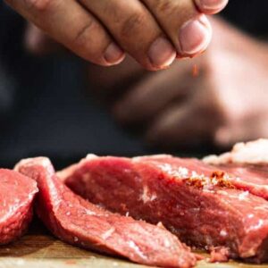 carne para churrasco: mão de pessoa colocando temperos em cortes de carne que estão na mesa