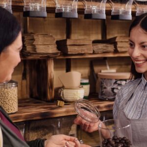 Cheerful shopkeeper helping customer in packaging free shop. Zero waste shopping - woman buying healthy chocolate in package free store.