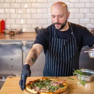 economia 2023: pizzaiolo com avental e luvas na cozinha terminando de rechear uma pizza em cima da bancada