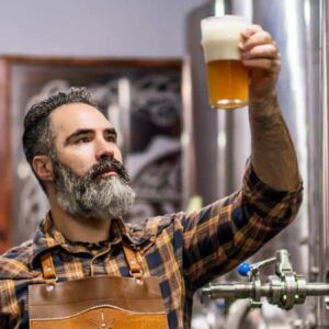 microcervejaria: empreendedor cervejeiro usando avental dentro de sua fábrica segurando copo de cerveja na mão