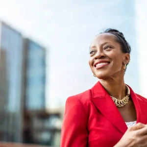 Mulher sorridente usando blazer vermelho em ambiente urbano moderno, segurando um smartphone na mão e olhando para o lado, com fundo desfocado de prédios
