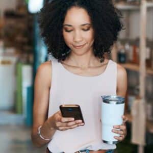 pesquisa de satisfação: mulher andando enquanto olha para celular e segurando caneca térmica