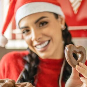 promoção de natal: mulher com gorro de natal e blusa vermelha sorrindo enquanto segura biscoitos de chocolate em formato de coração