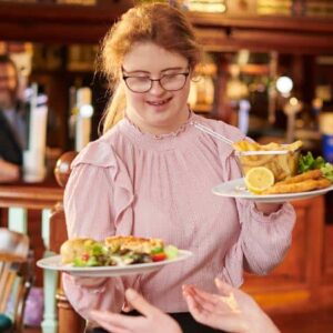 sazonalidade: garçonete com síndrome de down entregando pratos de comida para duas clientes diversas sentadas em mesa de restaurante