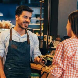 tributo vinculado: cliente sendo atendida no balcão por funcionário de cafeteria para fazer o pagamento enquanto existe outro funcionário trabalhando nos fundos e a amiga da cliente espera do lado